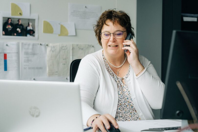 Frau mit Brille und weissem Cardigan sitzt am Schreibtisch im Büro, telefoniert mit dem Smartphone und arbeitet gleichzeitig am Computer. Im Hintergrund sind Notizen und Fotos an der Wand befestigt.