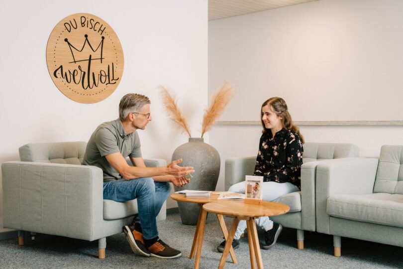 Ein Herr und eine junge Dame führen sich gegenüber sitzend ein Gespräch. Beide sitzen auf Sesseln. Zwei hölzerne Beistelltische trennen Sie voneinander. In der Ecke ist eine grosse Vase mit Pappeln und an der linken Wand ein Holzschild worauf steht: "Du bisch wertvoll"